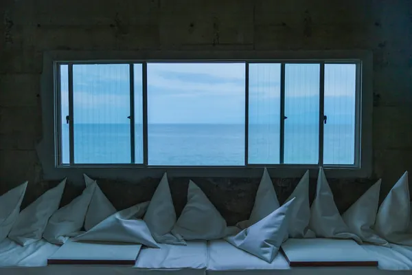 Banquette de coussins blancs devant une longue fenêtre ouverte sur la mer